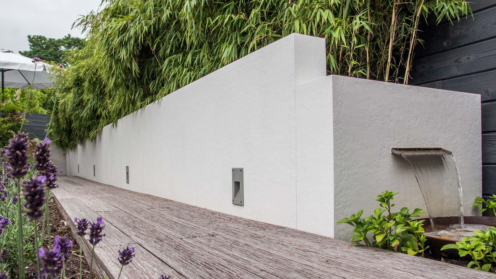Modernes Gartengestaltungselement mit weißer Wand, integriertem Wasserfall, umgeben von Bambuspflanzen und Holzboden im Vordergrund.