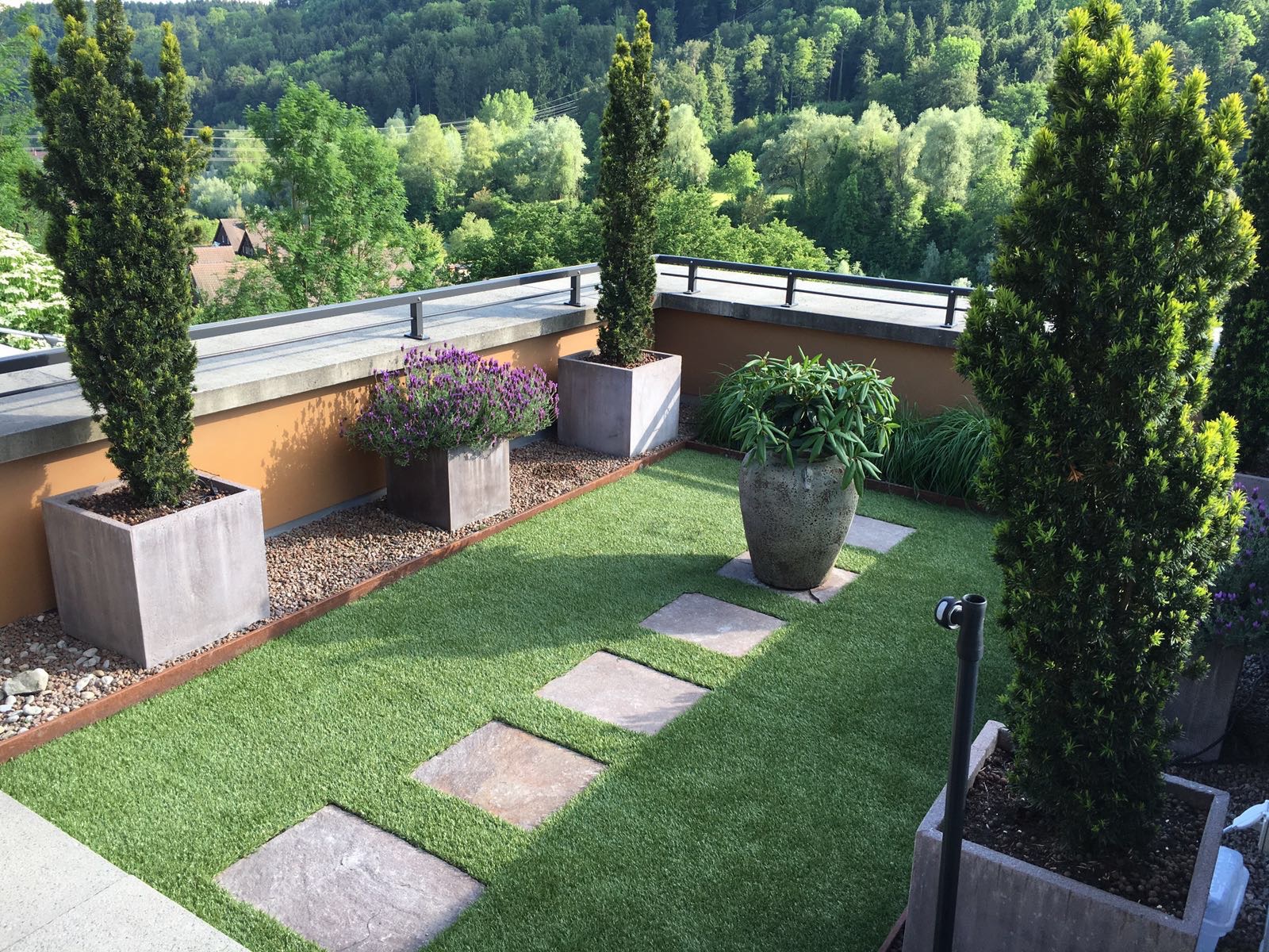 Gartenideen für Dachterrassen mit kunstvollem Pflasterweg, verschiedenen Pflanzen in großen Töpfen und einem Ausblick auf bewaldete Hügel.